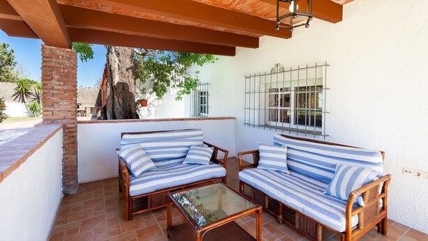 Une terrasse couverte avec deux canapés à rayures, une table basse en verre et un luminaire suspendu.