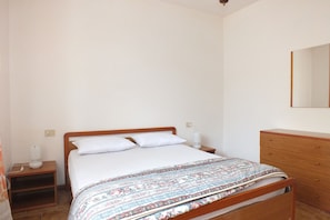 A bedroom with a wooden bed, white bedding, a nightstand with a lamp, and a dresser.