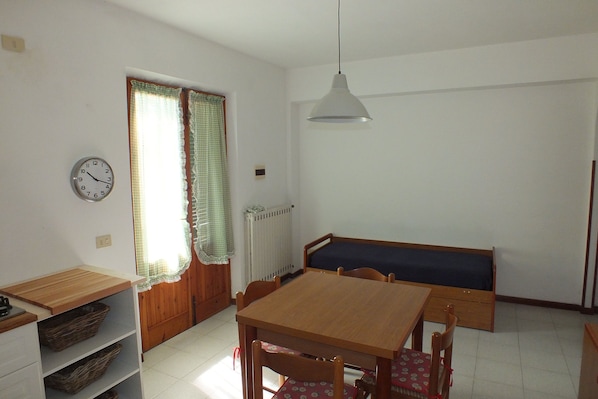 A dining area with a wooden table, chairs, a bench, a hanging light fixture, a clock, and a window with curtains.