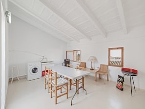 A white-walled room with a table, chairs, a washing machine, a lamp, and a grill.