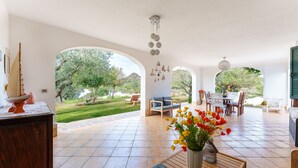 A spacious patio with a dining area, a wooden table with a vase of flowers, and a view of a lush garden. - Rustic Villa, Air Conditioning and Beautiful Garden (Orosei)