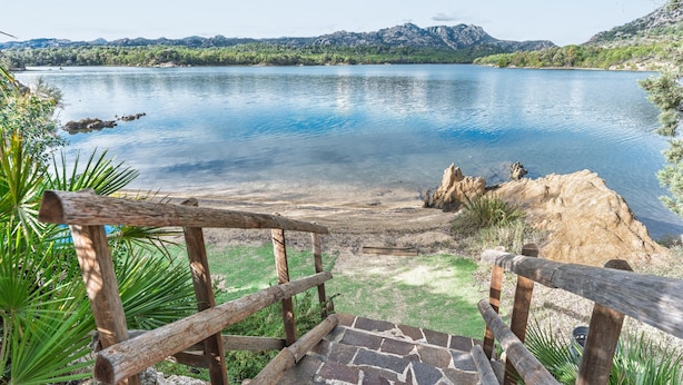 A wooden deck with a stone pathway leading to a lake with clear water and mountains in the background. - Rustic Villa, Air Conditioning and Beautiful Garden (Orosei)