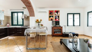 A kitchen with a dining table, chairs, and a TV. There is a window with curtains, a brick wall, and artwork on the wall. - Rustic Villa, Air Conditioning and Beautiful Garden (Orosei)