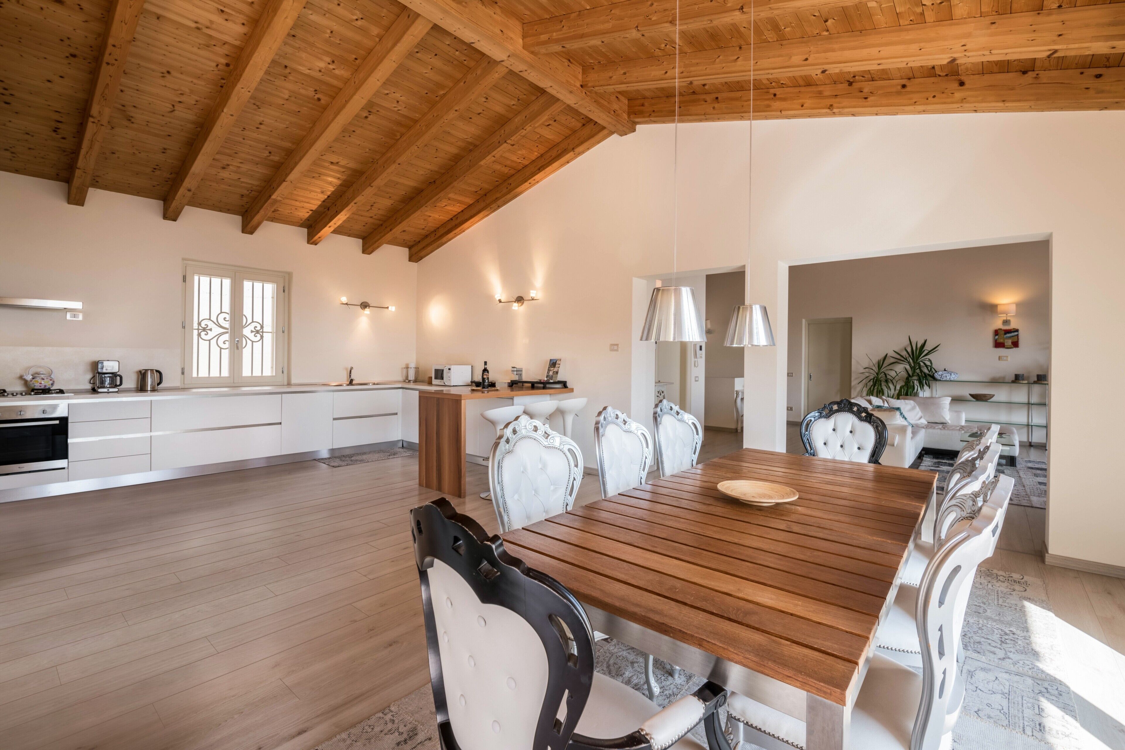Une cuisine spacieuse avec des poutres en bois, un coin repas avec une table en bois et des chaises blanches, et un îlot de cuisine moderne.