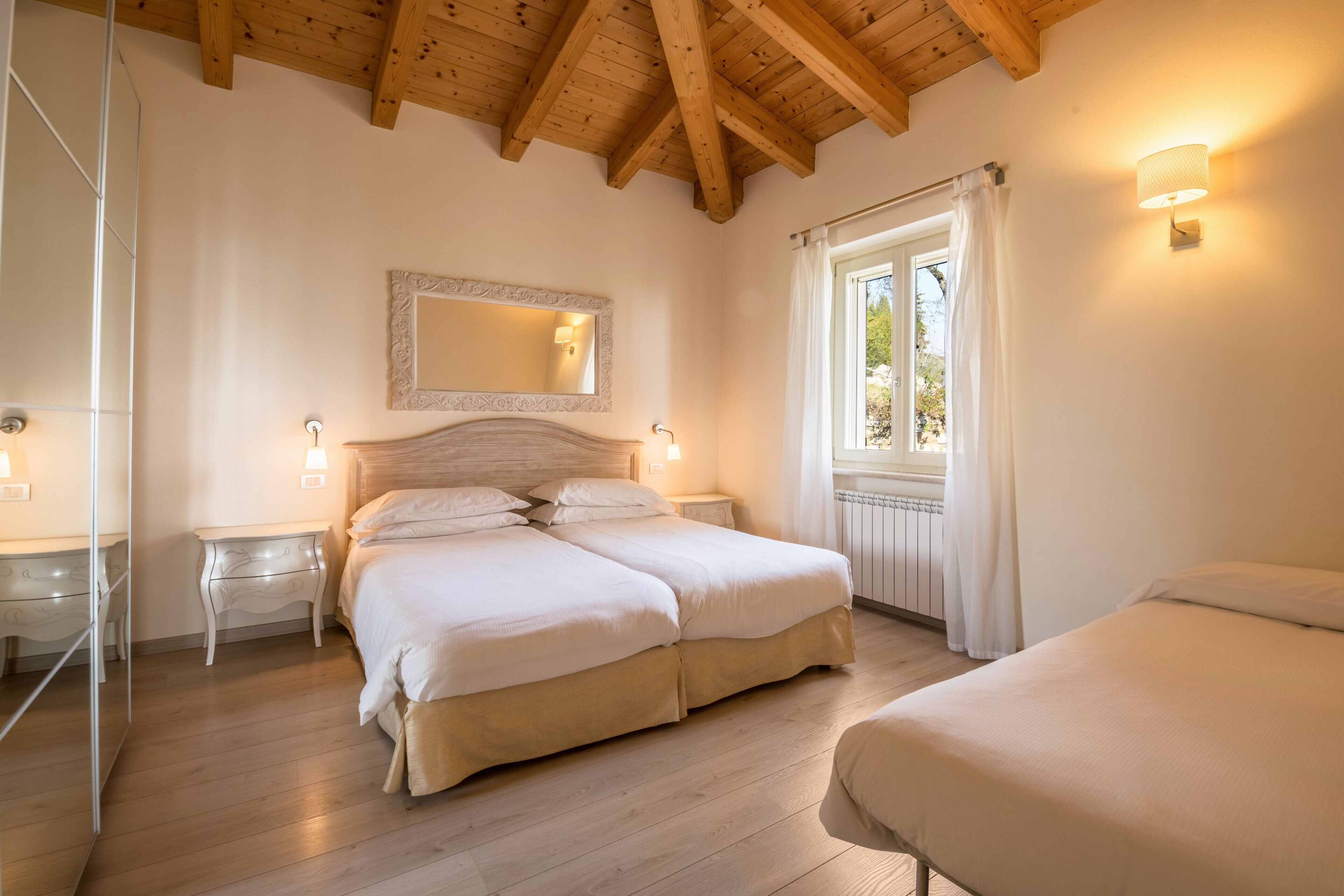 Une chambre avec un plafond en bois, un grand lit, des tables de chevet, un miroir et une fenêtre avec des rideaux.