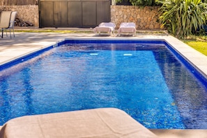 A rectangular swimming pool with a stone wall and a covered seating area with lounge chairs.