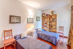 Un dormitorio con dos camas, una mesita de noche con lámpara, una estantería y una silla.