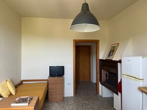 A bedroom with a bed, a wooden dresser, a television, a refrigerator, and a hallway leading to another room.