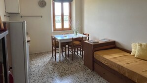 A compact room with a kitchenette, a dining table with chairs, a window with a view, and a bed with a pillow.