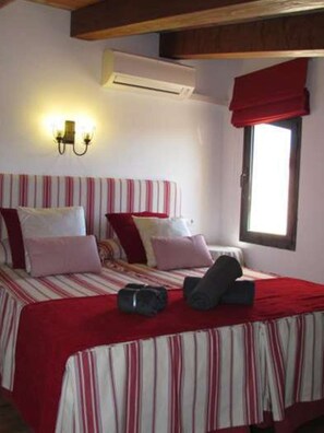 A bedroom with a bed, pillows, a red blanket, a window with a red valance, and an air conditioning unit.