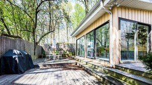 A wooden deck with a covered grill area, a table and chairs, and a building with large windows. - UNION PIER HOME WITH PRIVATE HEATED POOL and BEACH ACCESS! (Lakeside)