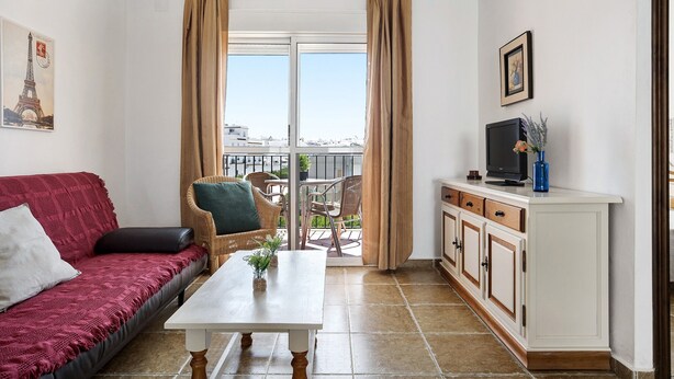 A room with a red sofa, a white table, a TV on a cabinet, and a balcony with a view.