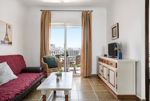 Une pièce avec un canapé rouge, une table blanche, un téléviseur posé sur un meuble et un balcon avec vue.