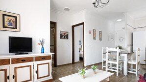 A living room with a TV, white furniture, a dining area, and a hallway.