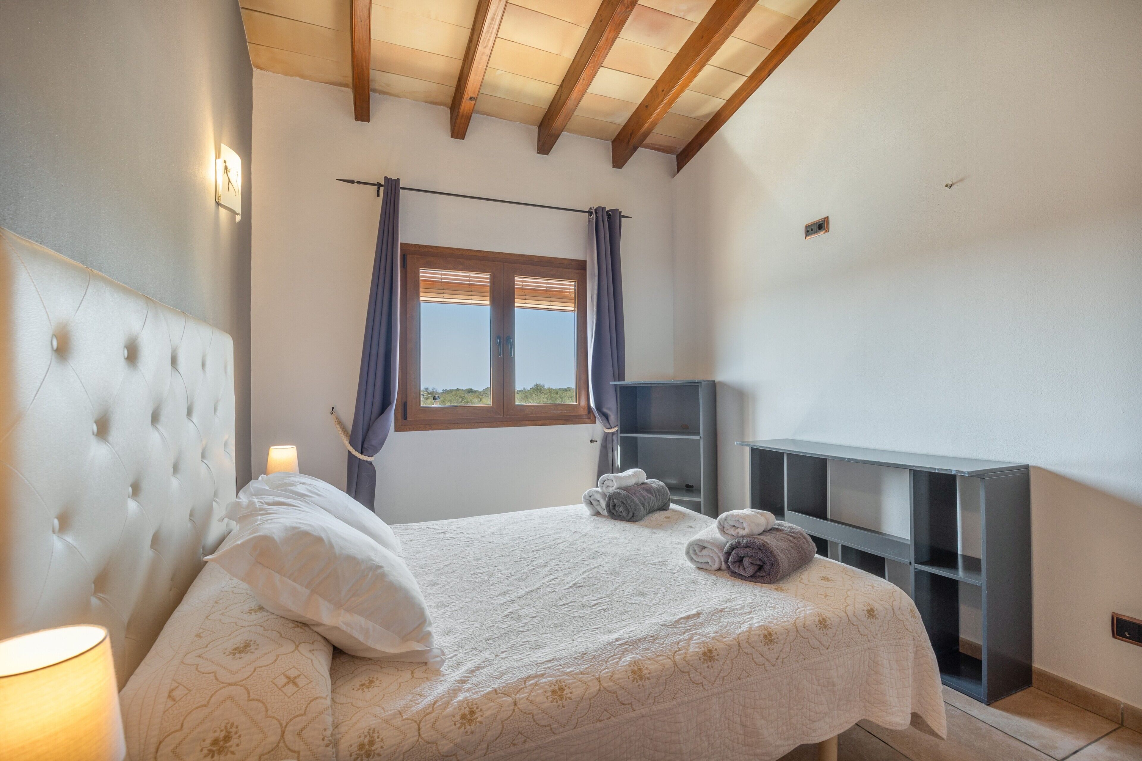 A bedroom with a bed, a nightstand, a lamp, a window with curtains, and a built-in TV shelf.