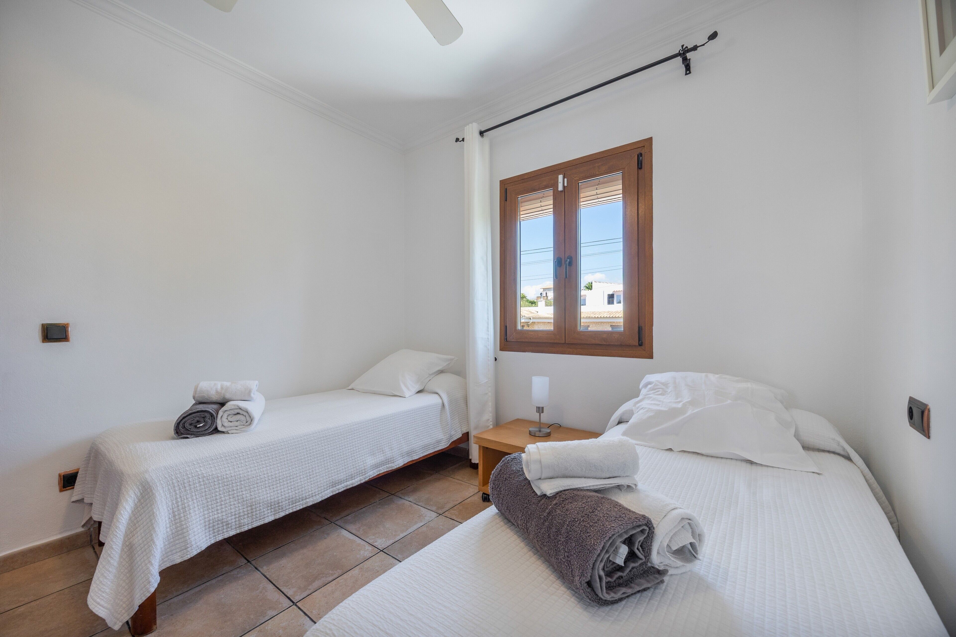 A small, clean room with two single beds, a bedside table with a lamp, and a window with wooden shutters.