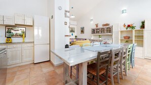 Een keuken met witte kastjes, een eettafel met stoelen en een koelkast.
