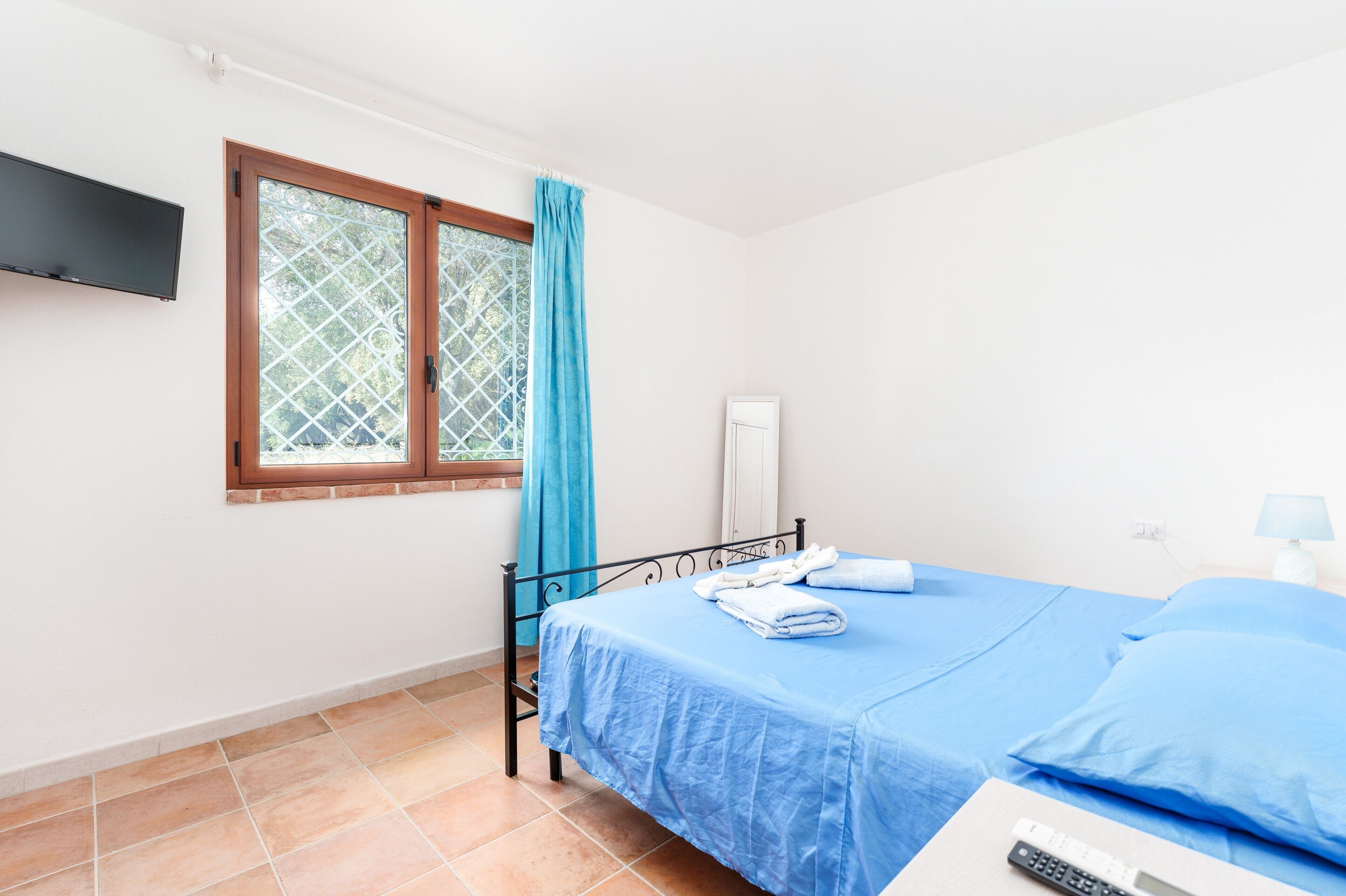 Een slaapkamer met een bed, een aan de muur bevestigde televisie, een raam met gordijnen en een nachtkastje met een lamp.
