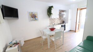 Une cuisine moderne avec des meubles blancs, une table à manger avec des chaises, un téléviseur à écran plat fixé au mur et un canapé vert dans l’espace salon.