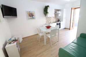 A modern kitchen with white cabinets, a dining table with chairs, a flat-screen TV mounted on the wall, and a green sofa in the living area.