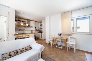 A modern kitchen with white cabinets, a dining area with a wooden table and chairs, and a white sofa with a patterned pillow.