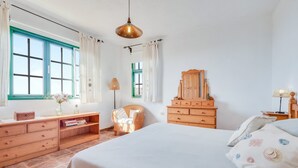 Een slaapkamer met een bed, een houten dressoir, een rieten stoel en een houten kast.