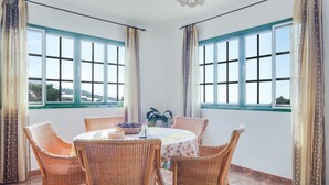 Een kamer met een ronde tafel, rieten stoelen en grote ramen voorzien van gordijnen.