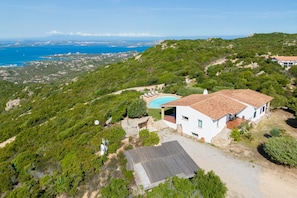 A white villa with a swimming pool, surrounded by lush greenery and overlooking a coastal view.