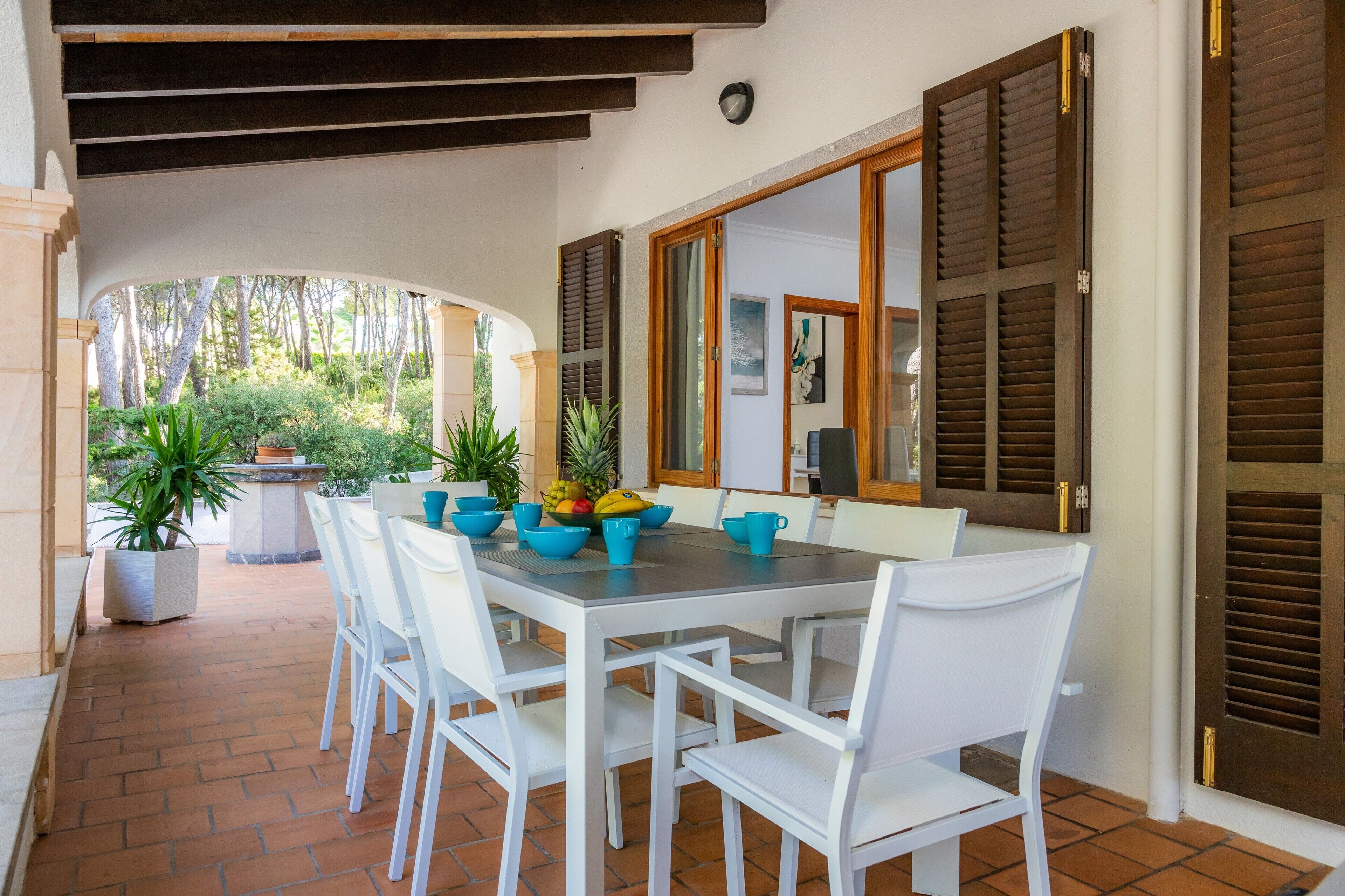 Um pátio externo com mesa e cadeiras, plantas em vasos e vista para o verde ao redor.