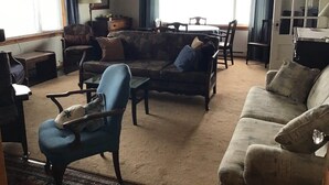 Una sala de estar con sofá, sillones, una mesa de centro y una mesa de comedor. Se ve una ventana con cortinas, una lámpara y una puerta con espejo.