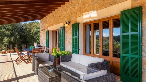 Un espacio de estar techado al aire libre con mobiliario de mimbre, contraventanas verdes y una pared de piedra.