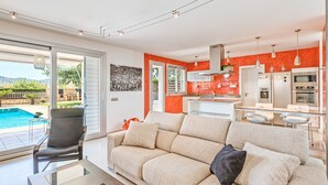 Een moderne woonkamer met een bank, een glazen salontafel en een stoel. Er is een keuken met een eettafel en stoelen, en een groot raam met uitzicht op een zwembad.