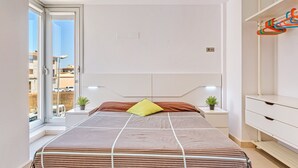 Een moderne slaapkamer met een groot bed, een raam met uitzicht op gebouwen, een nachtkastje met een plant en een witte kledingkast.
