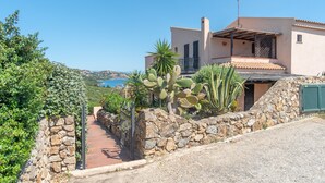 A Mediterranean-style villa with a stone wall, cactus plants, and a view of the sea.