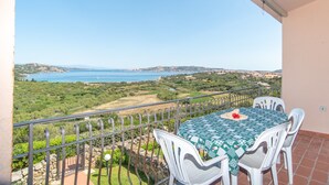 A balcony with a table and chairs, overlooking a coastal landscape with greenery and a view of the sea.