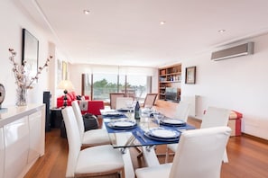 Un espace repas moderne avec une table en verre pour six personnes, des chaises blanches, un canapé rouge et un coin cuisine doté de meubles blancs et d’une chaise rouge.