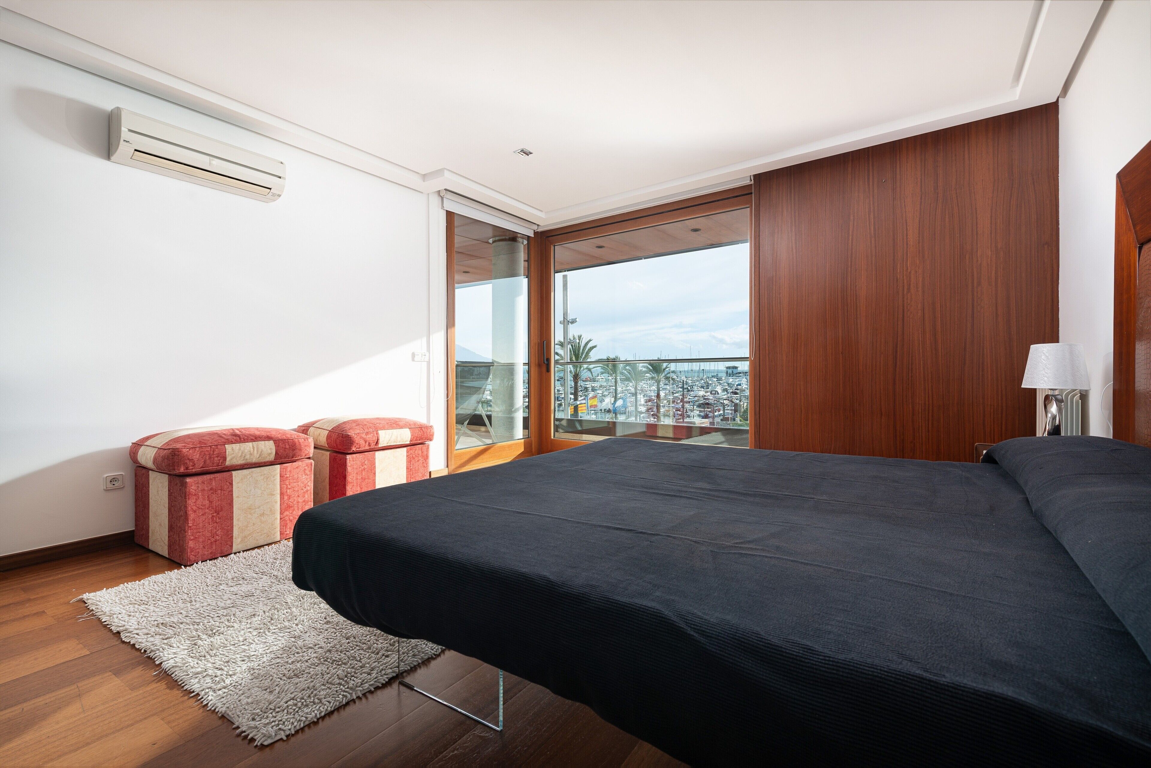 Un dormitorio con una cama, un reposapiés rojo y beige, una alfombra blanca y vistas a un balcón con barandilla.