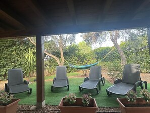 Een overdekt terras met vier loungestoelen en een hangmat, omgeven door groen en bomen.