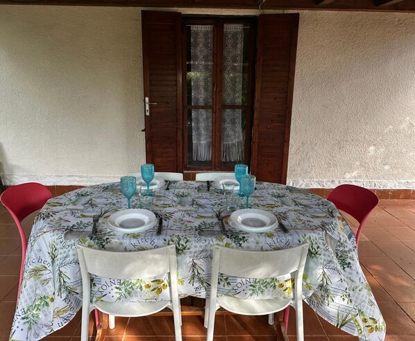 Une table ronde recouverte d’une nappe à motifs floraux, avec des chaises blanches et de la verrerie bleue, le tout disposé pour quatre personnes.