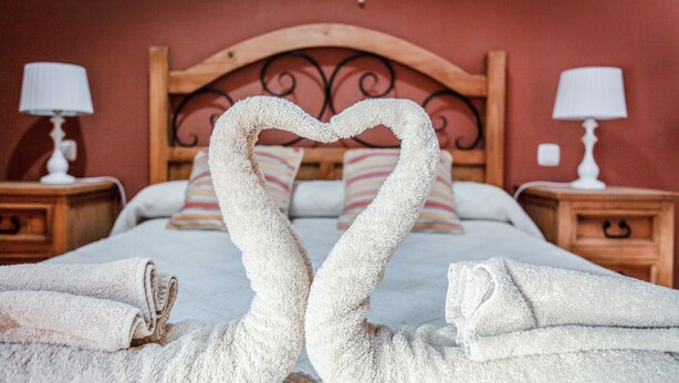 Een bed met een handdoekarrangement in de vorm van een hart, een houten hoofdbord, gestreepte kussens en bedlampjes.