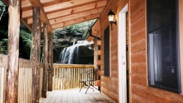 A wooden cabin with a wooden deck, a waterfall view, and a single chair.