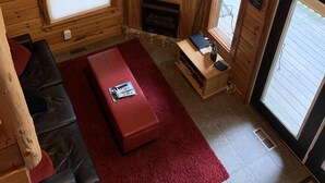 A cozy cabin interior with a wooden ceiling, a television on a wooden stand, a fireplace, a red sofa, and a window with blinds.
