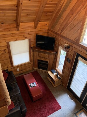 L’intérieur d’un chalet accueillant, avec un plafond en bois, un téléviseur posé sur un meuble en bois, une cheminée, un canapé rouge et une fenêtre avec des stores.