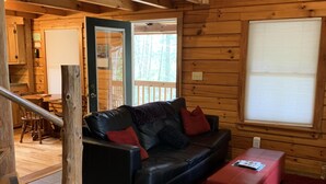 A log cabin interior with a black leather sofa, red cushions, a red rug, a wooden staircase, and a window with blinds.