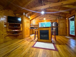 Interior de una cabaña de madera con chimenea, pisos de madera, una zona de cocina y un televisor.