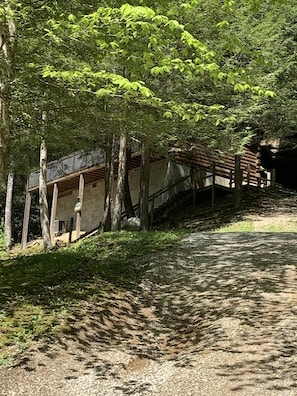 A rustic cabin with a wooden deck and a stone wall, surrounded by dense greenery.