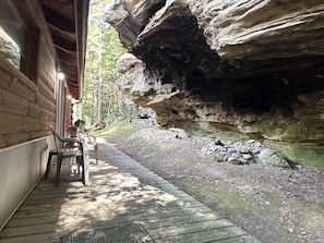 A wooden deck with chairs, a table, and a view of a rocky cliff and forest. - Cozy secluded cabin with rock overhand and Wi-Fi (Rogers)