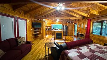 A cabin interior with a wooden ceiling, a sofa, a bed, a TV, a kitchen area, and a fireplace.