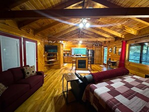 A cabin interior with a wooden ceiling, a sofa, a bed, a TV, a kitchen area, and a fireplace. - Cozy secluded cabin with rock overhand and Wi-Fi (Rogers)