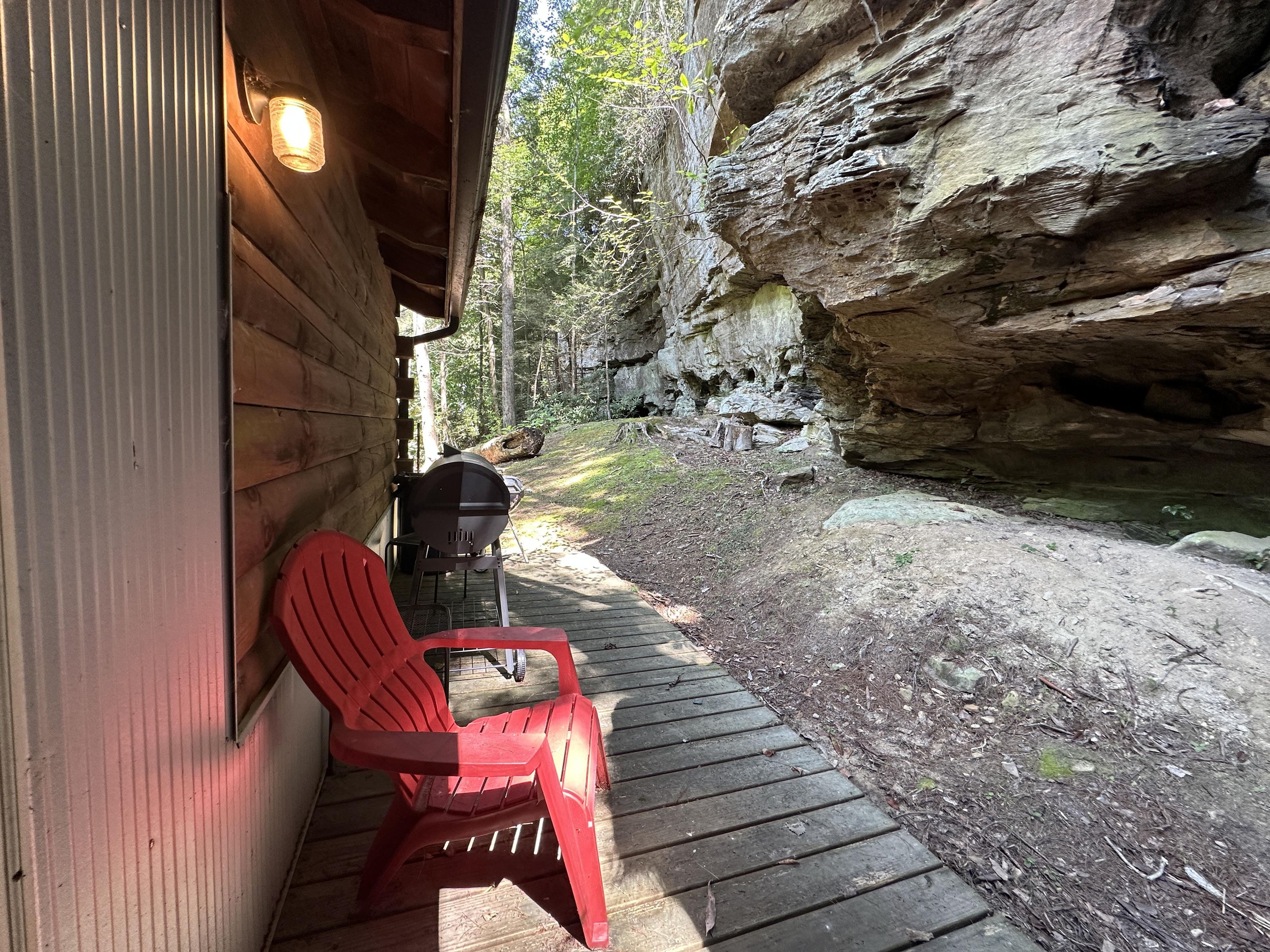 A wooden deck with a grill and red chairs, a rock wall, and a wooden cabin.